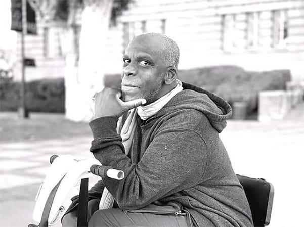 Leroy F. Moore Jr. seated in a wheelchair, wearing a hooded sweatshirt and scarf, and looking toward the camera with one hand near his chin. Black-and-white photograph with a softly blurred urban background.